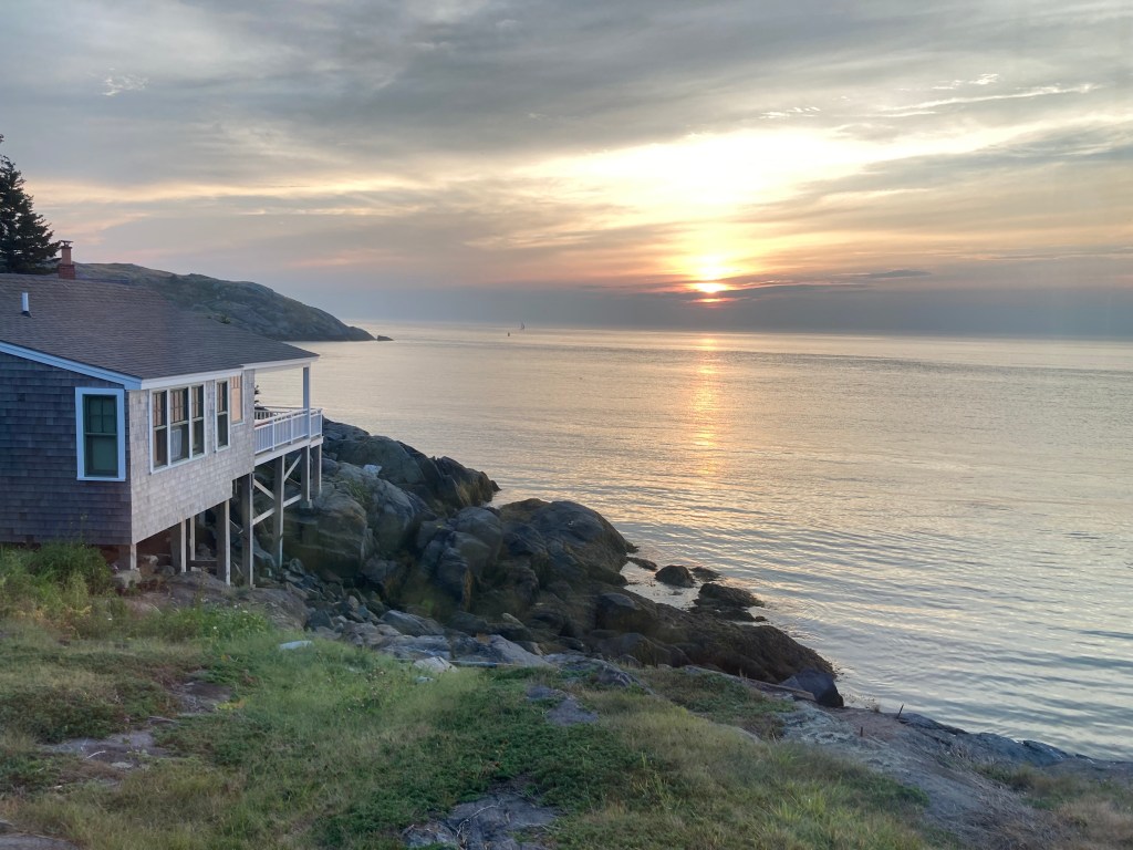 A golden sunset over a calm ocean. The shore is rocky and a gray house on stilts is to the left.