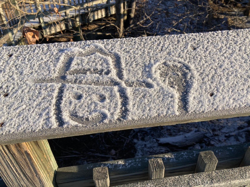 A snow-covered fance railing with a smiley face drawn in the snow. The face is topped by a flat-brimmed hat like park rangers wear.